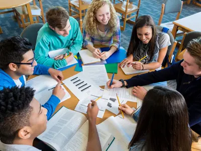 A group of smiling teens sitting around a table covered in homework.
