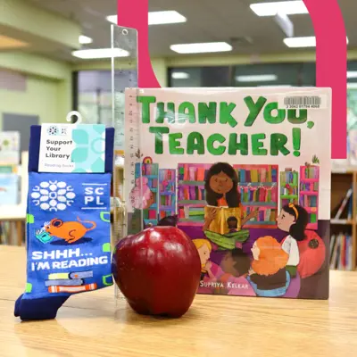 A pair of socks are posed with a red apple, ruler, and book titled "Thank You, Teacher".