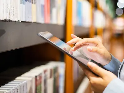 Hands holding a tablet beside a bookcase.