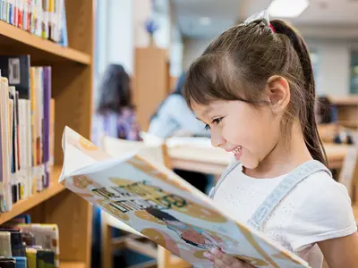 A young girl smiling while reading a book at the library.
